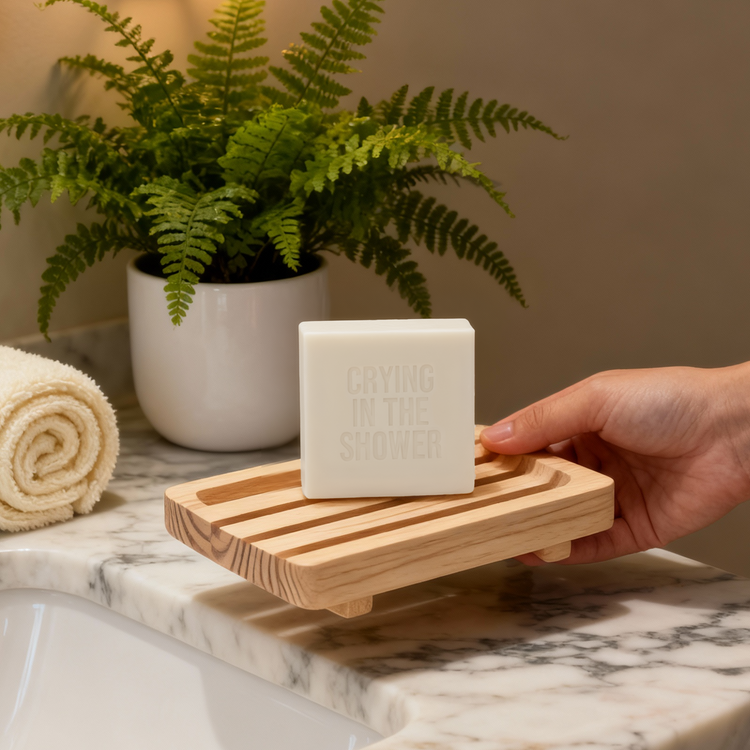 Hand holding a bar of Crying in the Shower soap on a wooden soap dish with a plant and towel in the background.